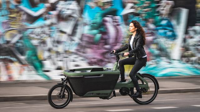 Frau in Lederjacke fährt ein grünes Lovens-Lastenrad vor einer Graffiti-Wand, dynamisches Stadtszenario