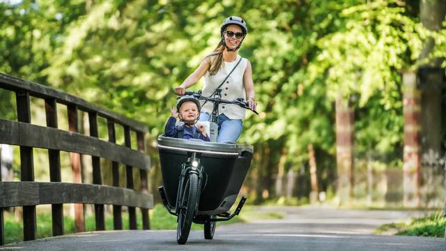 rau fährt ein Lastenrad auf einem Waldweg, Kind sitzt in der Transportbox und winkt fröhlich in die Kamera